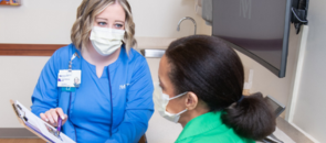 A female nurse in blue scrubs holding and pointing to a clipboard while talking to a patient.